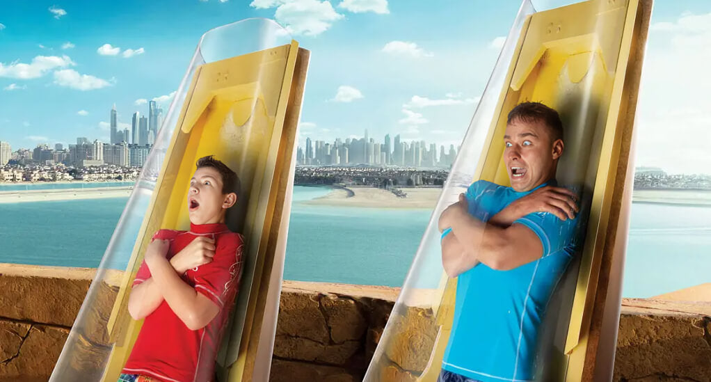 Two thrill-seekers bracing for the vertical drop on Poseidon's Revenge slide at Atlantis Aquaventure Waterpark, with Dubai skyline in the background.