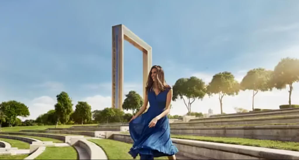 Woman in a flowing blue dress posing near the iconic Dubai Frame in Zabeel Park on a sunny day.