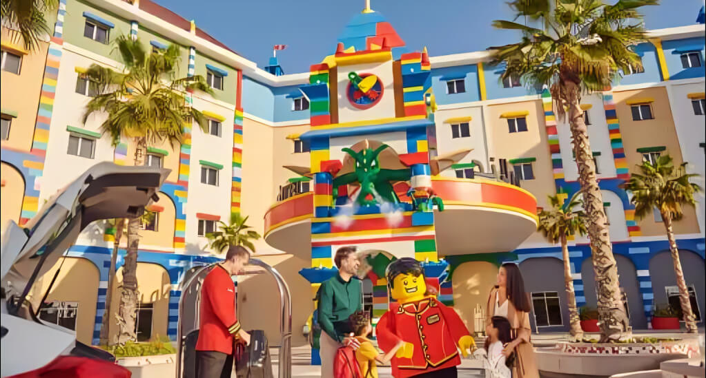 Family arriving at the colorful LEGO-themed hotel at LEGOLAND Dubai, greeted by a life-sized LEGO bellhop figure and resort staff.