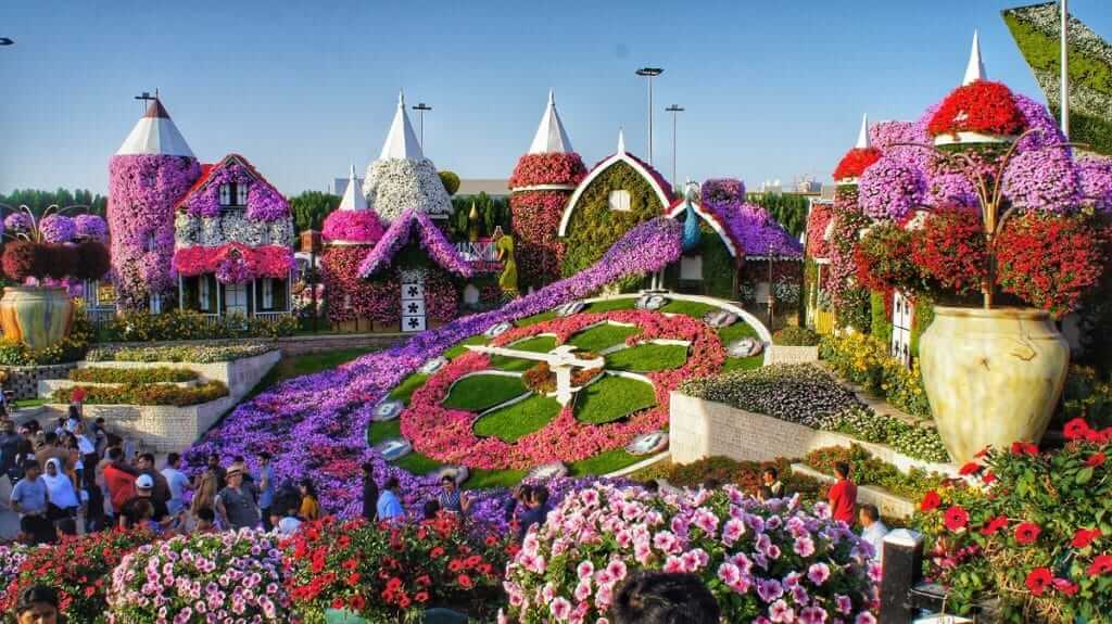 Stunning floral clock surrounded by flower-covered houses and vibrant blooms at Dubai Miracle Garden, with visitors enjoying the colorful landscape.