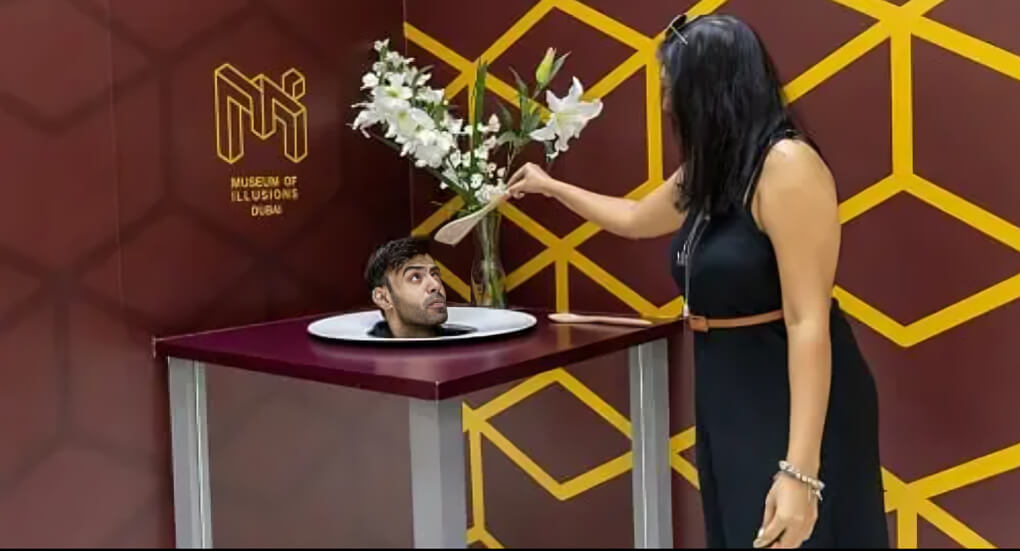 Interactive optical illusion exhibit at the Museum of Illusions Dubai, where a guest playfully appears to serve a man's head on a platter, creating a mind-bending visual trick.